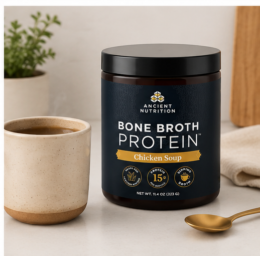 A jar of Ancient Nutrition Bone Broth Protein in Chicken Soup flavor, styled on a light beige surface next to a ceramic cup, a gold spoon, and a small potted plant.