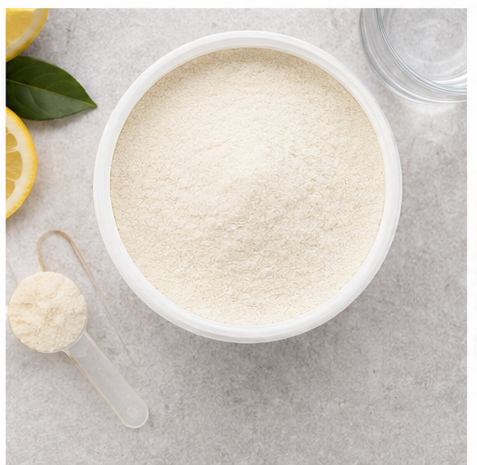 A white bowl filled with a fine off-white powder, shown from above on a light gray surface with a small clear scoop, a glass jar edge, lemon slices, and green leaves around it.